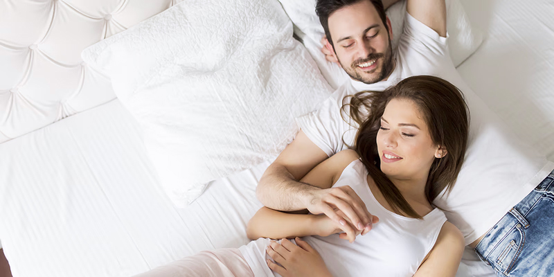 Ζευγάρι ξαπλωμένο σε ένα κρεβάτι, αγκαλιασμένο και χαμογελαστό. Ο άνδρας βρίσκεται ξαπλωμένος στο πλάι, ενώ η γυναίκα είναι ξαπλωμένη μπροστά του, με το κεφάλι της να στηρίζεται στο στήθος του. Και οι δύο φορούν άνετα, ανοιχτόχρωμα ρούχα και δείχνουν χαλαροί και ευτυχισμένοι, απολαμβάνοντας τη στιγμή μαζί.